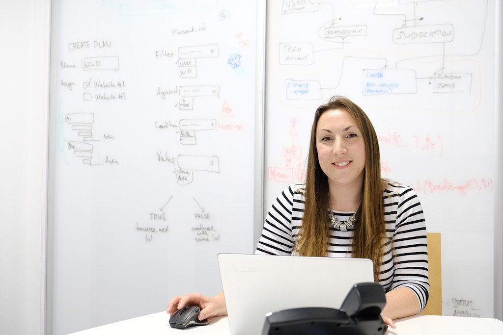 Image of our team member Laurie, sitting at a laptop smiling, in front of a white board.